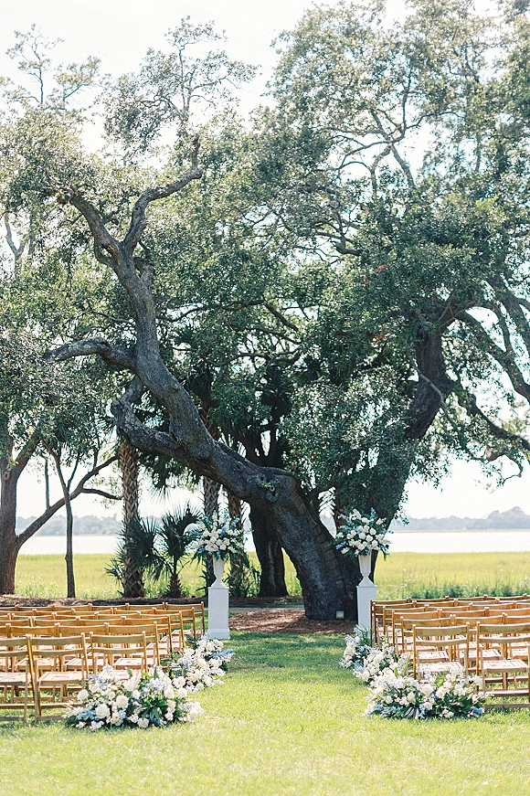 Outdoor ceremony setup with garden ceremony seating, wood folding chairs and white-green aisle florals beneath a large oak tree by the water view