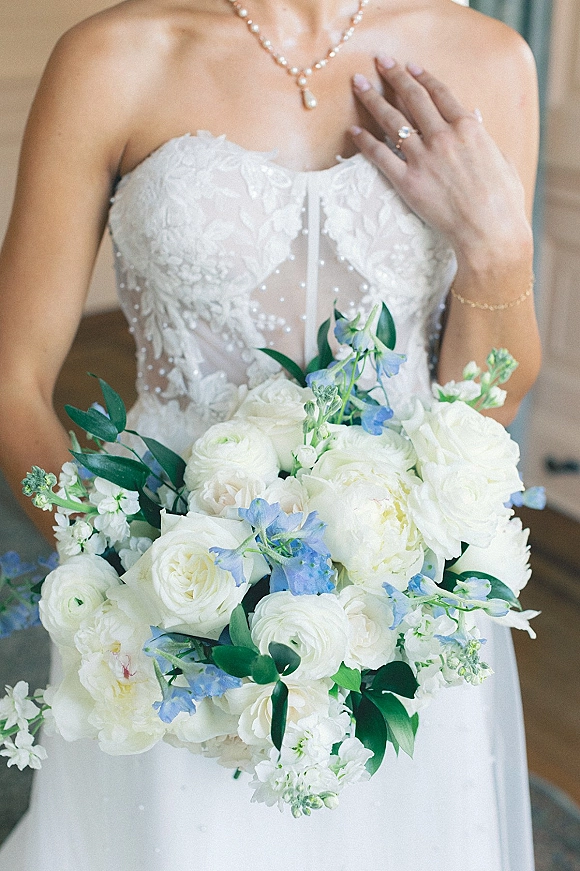 Bridal bouquet of white and blue wedding bouquet blooms with greenery held by a bride in a strapless lace gown in soft indoor light