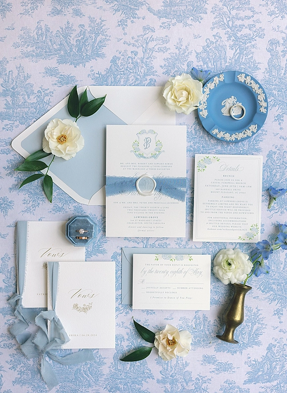Invitation suite with blue wedding invitations, wax-sealed envelopes, ribbon, rings on a blue plate, and flowers on blue toile fabric backdrop