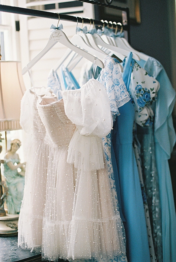 Bridesmaid dresses in blue bridesmaid dresses tones hang on bow ribbon hangers on a rack by a window, mixing floral and beaded styles