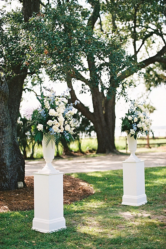 Ceremony aisle decor with wedding aisle pedestals holding white urn vases of white roses and blue blooms, lined on a sunlit oak path