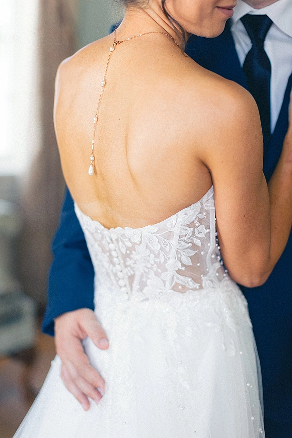 Wedding couple portrait of bride and groom embracing, highlighting her illusion lace back dress with pearl drop back necklace against a neutral wall