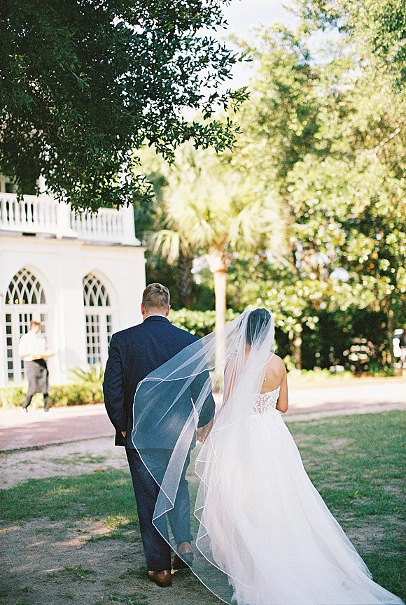 Bride and groom walk away holding hands, her long veil trailing over the lawn beside palm trees and a white building in back.