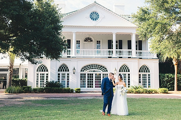 Couple portrait of bride holding bouquet as groom kisses her hand, veil flowing, in front of a white mansion with arched windows