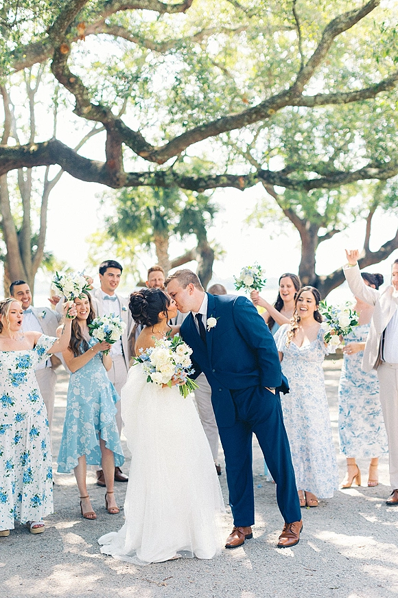 Wedding kiss as newlyweds kissing with cheering bridal party, bride in white gown and groom in navy suit on a tree-lined path