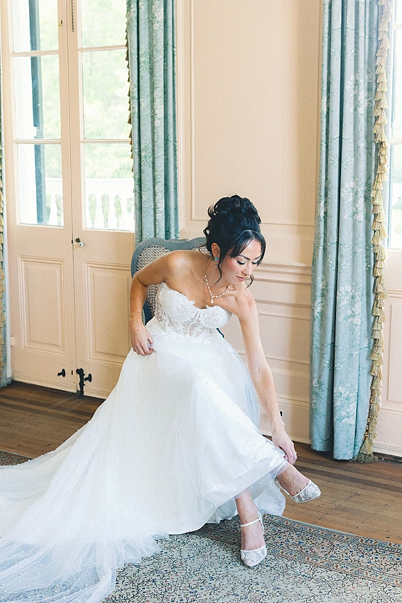 Bride getting ready as she puts on bridal heels beside a strapless lace gown with pearl necklace, framed by tall windows and paneled walls