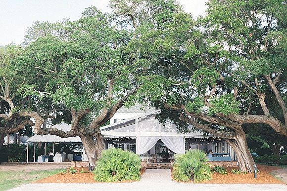 Outdoor wedding reception under a wedding reception tent with draped white fabric, string lights, and tables beneath oak trees