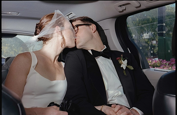 Wedding kiss as newlyweds in car backseat, bride in satin dress with veil and groom in tuxedo with glasses, street view outside window