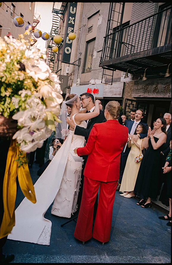 Wedding kiss as bride in a flowing veil and train embraces groom in black suit, guests cheering on a city street by brick facade