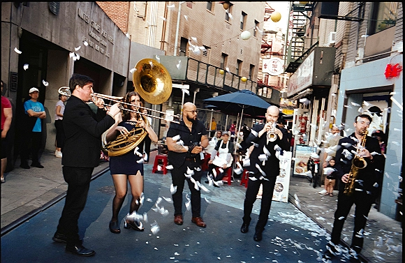 Wedding brass band leading a second line wedding down a brick-lined city alley, playing trombone and sousaphone as confetti falls