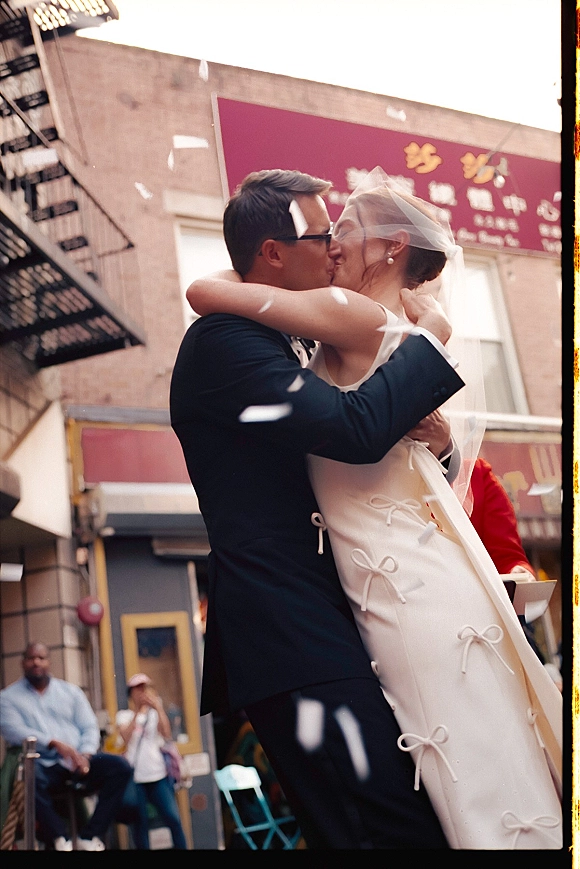 Wedding kiss as confetti falls around the bride in a veil and dress and groom in a tuxedo and eyeglasses on a city street by a brick building
