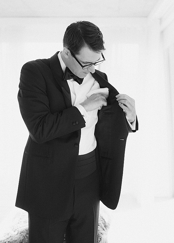 Groom getting ready in a black tuxedo, adjusting his pocket square with bow tie and eyeglasses in a simple indoor room with white wall