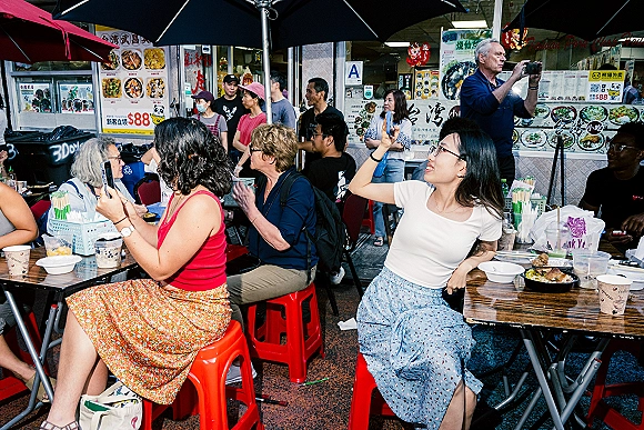 Street food dining at a night market food stall, with people eating at folding tables on plastic stools outside a menu-lined storefront.