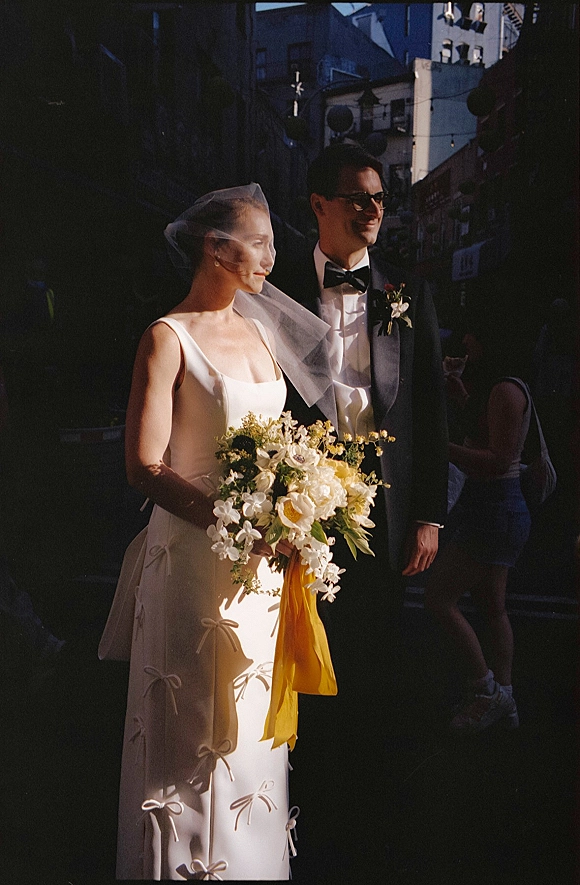 Couple portrait of bride holding a bouquet beside groom in tuxedo, her veil trailing on a sunlit city street with string lights