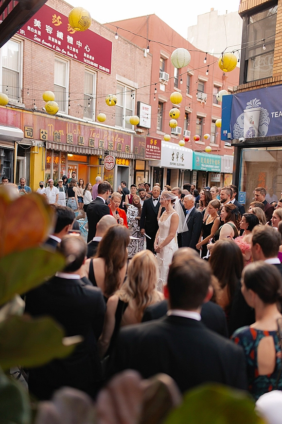 Wedding ceremony moment on a city street as bride in veil and groom in tuxedo face the officiant under string lights and lanterns