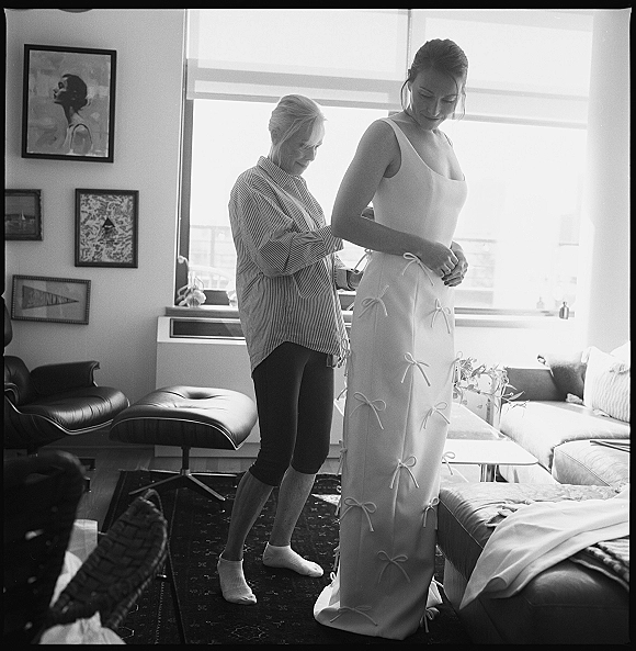 Wedding dress fitting as bride in a bow-detail bridal gown looks down while someone in a striped shirt adjusts it in a bright living room