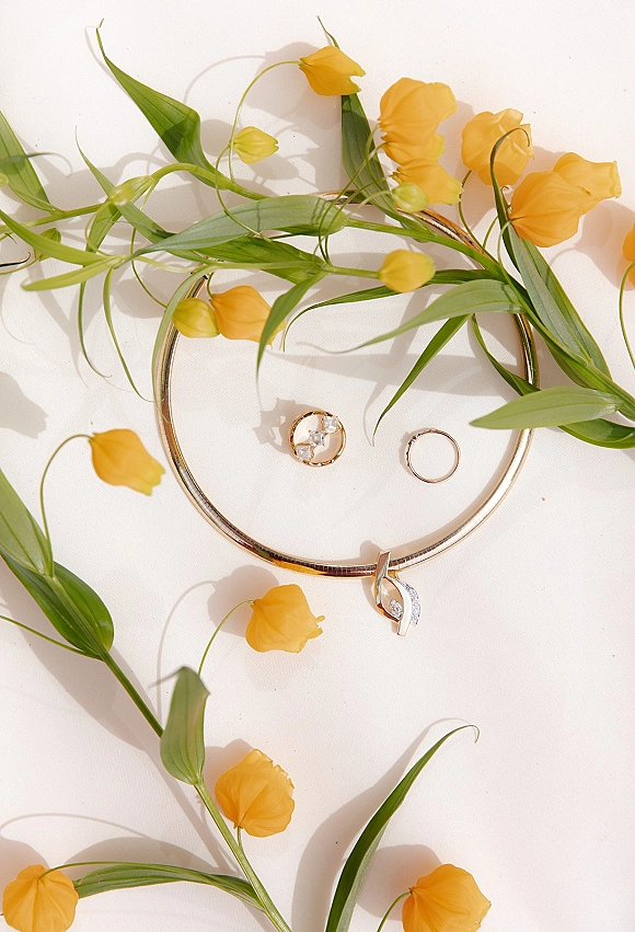 Wedding rings flatlay with gold bands and diamond ring beside necklace and earrings, accented by yellow flowers and green leaves on white surface