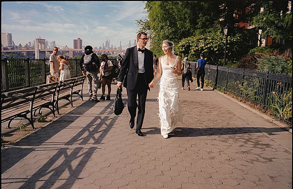 Wedding couple walking hand in hand, bride and groom holding hands with birdcage veil and mini bouquet on a park walkway by skyline and bridge