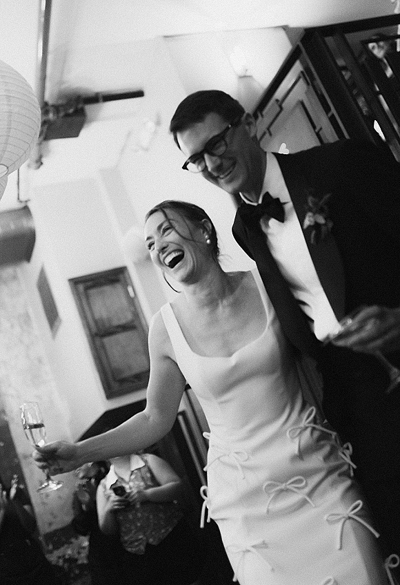 Reception moment as bride and groom laughing while entering, bride holding a champagne flute, guests and paper lanterns behind