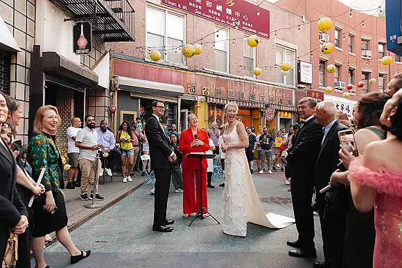 Wedding ceremony moment on a city street as the couple exchange vows at a stand with microphone under string lights and lanterns, guests around