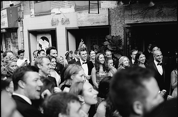 Wedding guests in formalwear laugh and cheer on a city street, tuxedos and evening dresses visible as some take smartphone photos