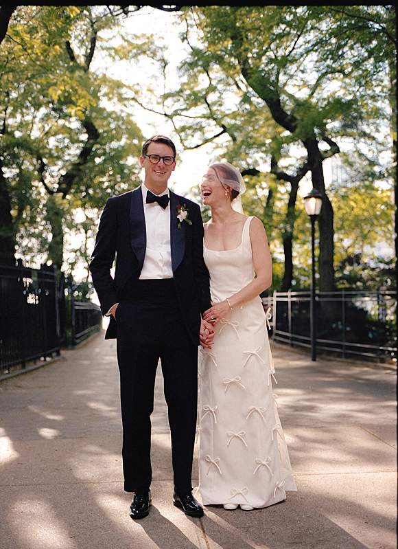 Couple portrait of bride and groom laughing, holding hands on a sunlit tree-lined park walkway, her veil flowing beside his tuxedo