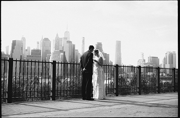 Couple portrait in a black and white wedding photo, bride in veil and groom in suit embracing by a waterfront fence with city skyline behind