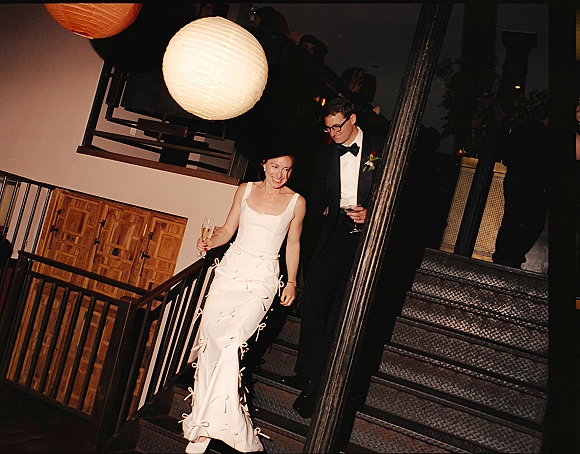 Wedding reception entrance as newlyweds enter on a staircase, bride in satin dress with champagne flute, groom in tux with cocktail glass, guests behind