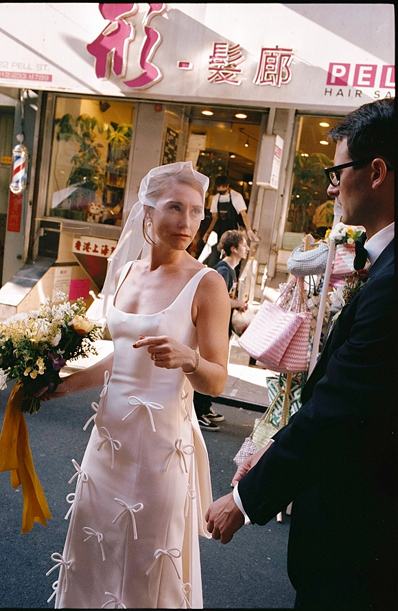 Couple portrait on a city street, bride with veil and colorful bouquet holding hands with groom in black tuxedo by storefront signage