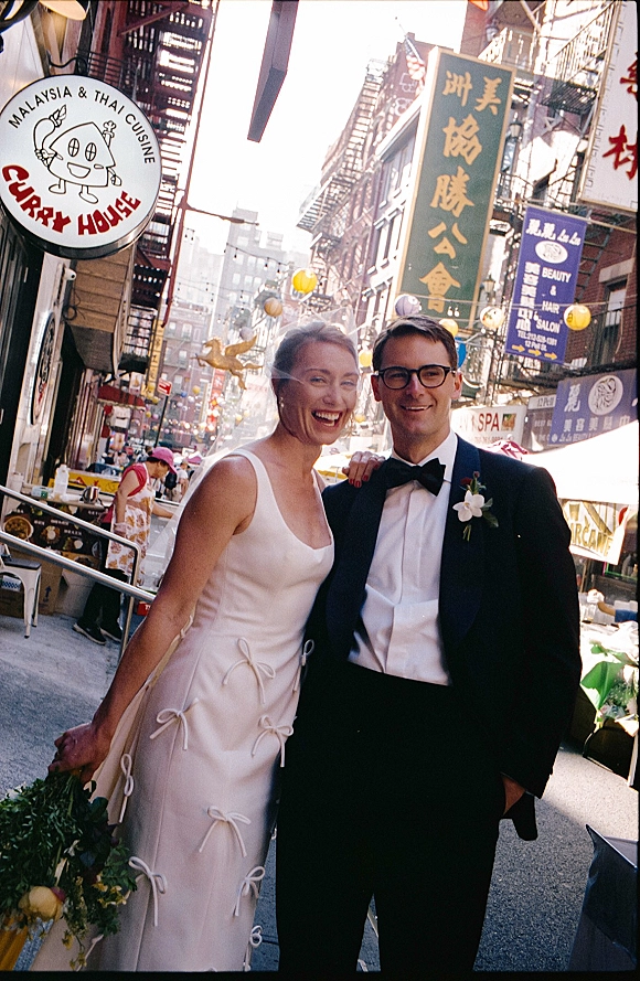 Couple portrait of bride in a satin wedding dress holding a bouquet and groom in tuxedo on a city street with lanterns and signs.