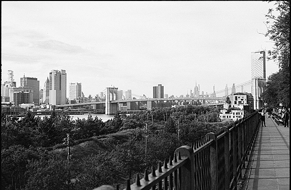 City skyline photo in black and white, viewed from a bridge walkway with railing and benches, overlooking a river and trees under open sky