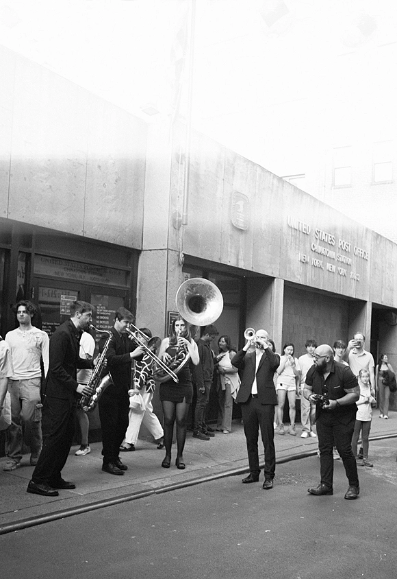 Wedding parade with a second line wedding brass band, trumpet and tuba leading suited guests and a dressed couple down a city street crowd