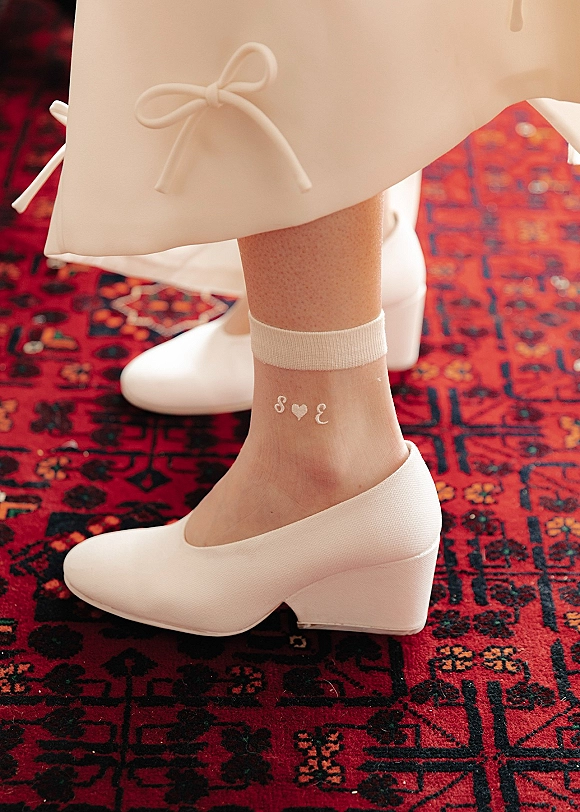 Bridal shoes, white wedge wedding shoes with monogram ankle socks and a heart, beneath a midi dress bow detail on a red patterned rug