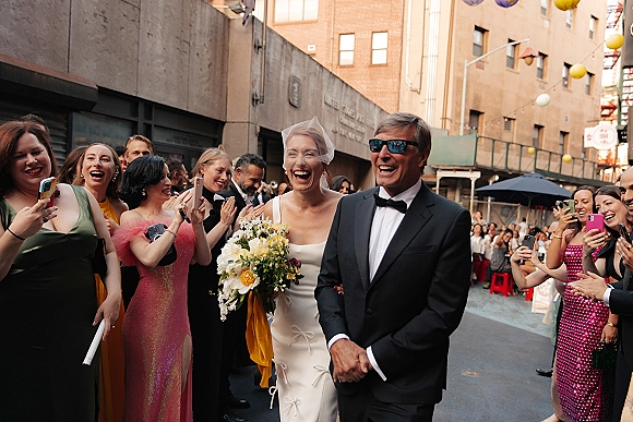 Wedding processional with bride walking down aisle in a white dress and veil, holding a bouquet, as guests cheer on a city street under lanterns