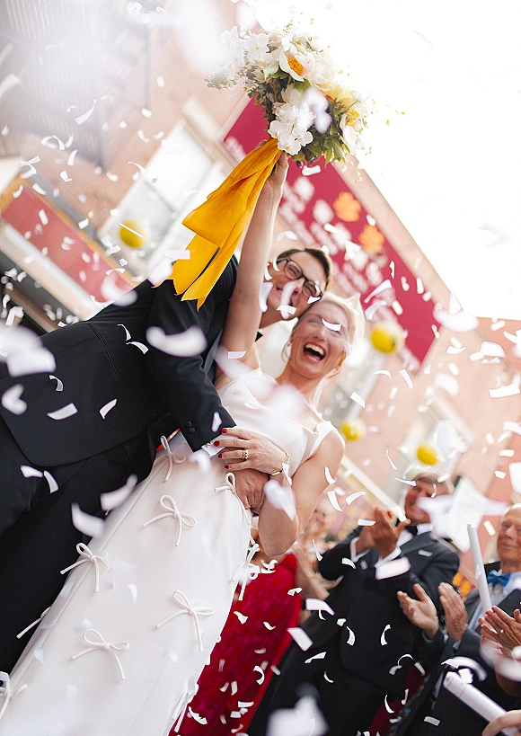 Wedding recessional with confetti wedding exit as the groom lifts the laughing bride, bouquet raised with yellow ribbon as guests cheer on a city street