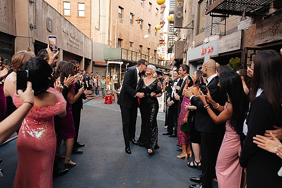 Wedding exit as groom in tuxedo kisses his mother in an evening gown and sunglasses, guests filming under string lights on a city street