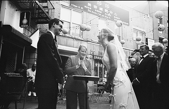 Wedding ceremony moment on a city street, couple exchanging vows as the bride in veil laughs under string lights with guests nearby