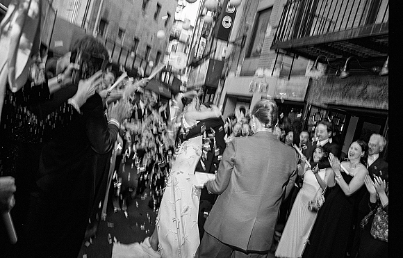 Wedding exit with confetti as bride in wedding dress and groom in suit walk down a city street while guests cheer and pop party poppers