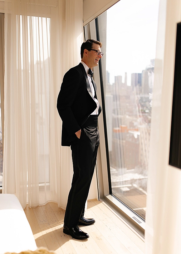 Groom portrait in a black tuxedo with bow tie and eyeglasses, standing by floor-to-ceiling windows with a city skyline view