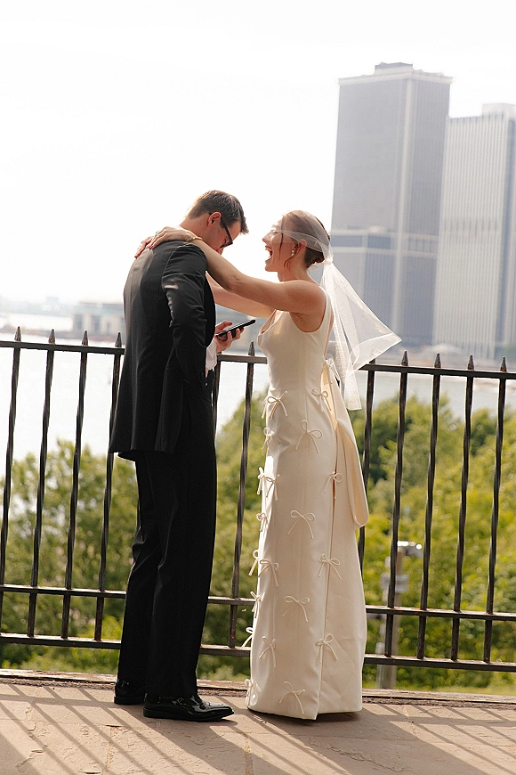 First look moment as bride taps groom’s shoulder on a terrace overlook, her veil lifting near iron railing with skyline and river beyond