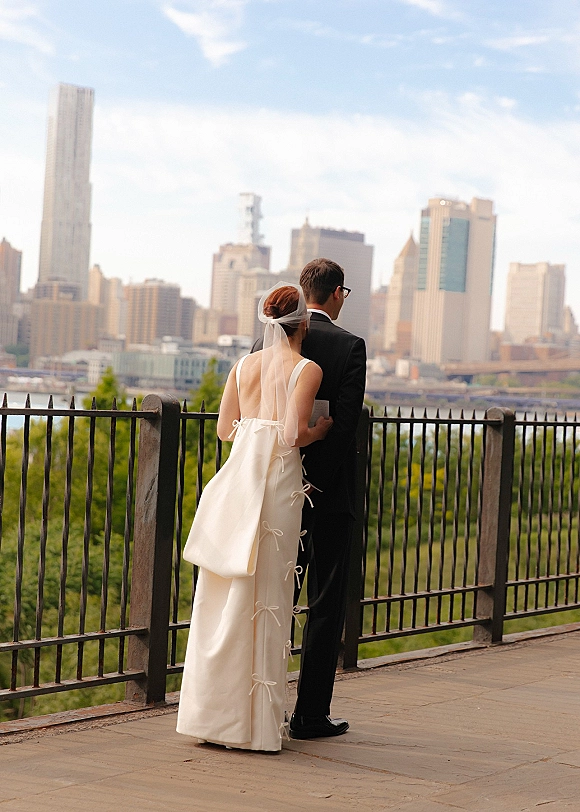 Couple portrait from behind of bride in veil and low-back dress and groom in tuxedo with glasses, overlooking a river city skyline
