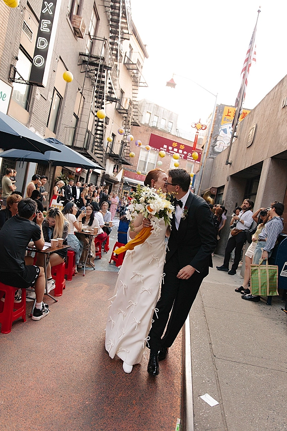 Wedding kiss portrait of bride and groom kissing while walking on a busy city sidewalk, bride holding bouquet beside cafe red stools