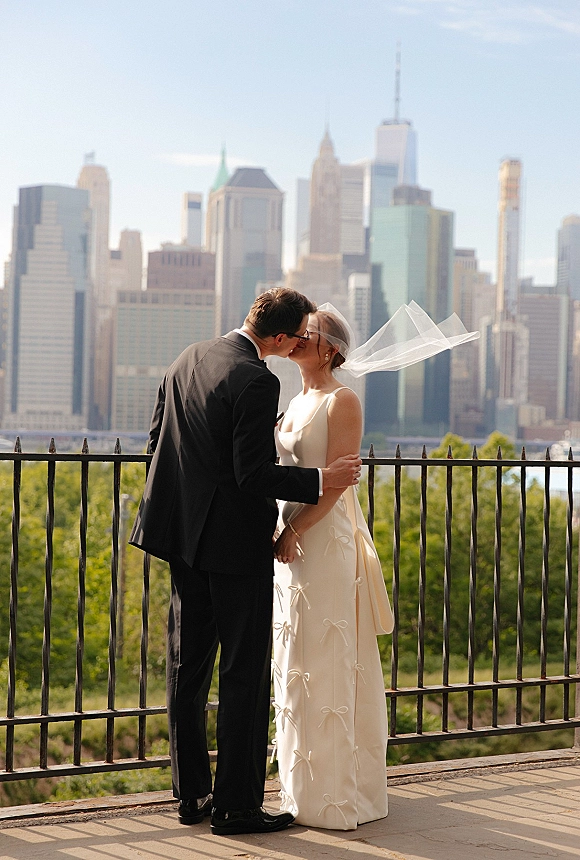Wedding kiss portrait of bride and groom kissing as her veil blows in the wind by an iron railing with city skyline and waterfront behind