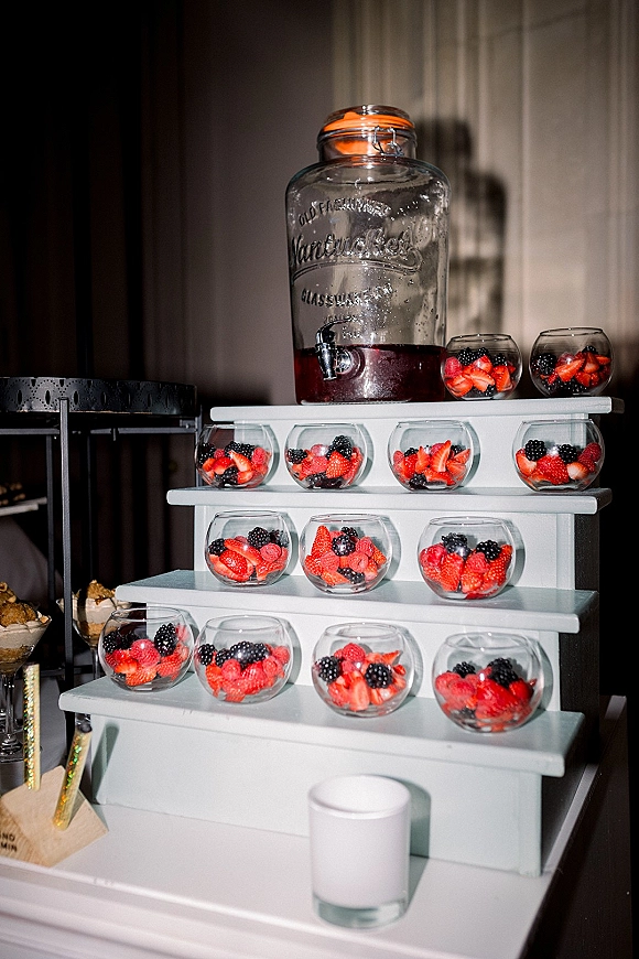 Dessert table with wedding dessert table berry dessert cups on a white tiered stand, plus glass drink dispenser, against curtain backdrop