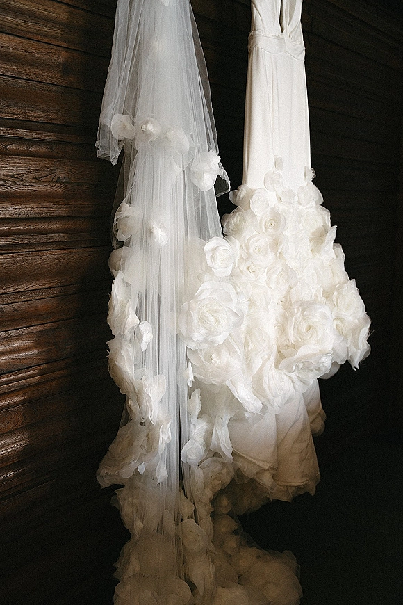 Wedding dress with 3D floral appliqué and a white tulle veil hanging against a rustic wood plank wall, showcasing textured bridal details