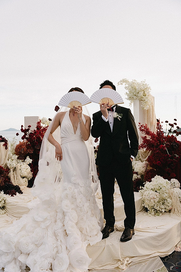 Couple portrait of bride and groom holding fans, she in halter ruffle dress and veil, he in tuxedo with rose boutonniere by water horizon