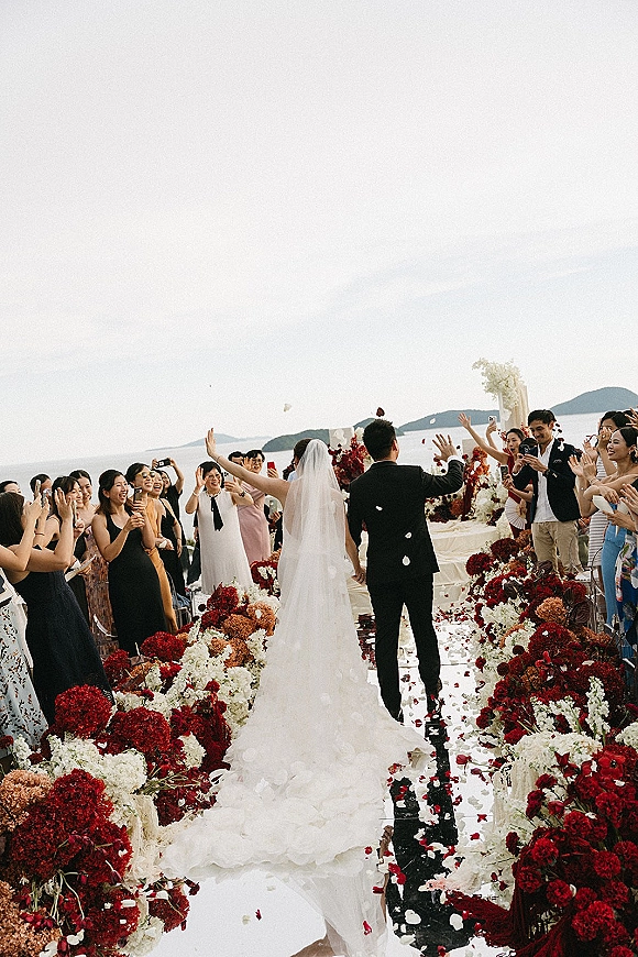 Wedding recessional as bride and groom walk away hand in hand down an outdoor wedding aisle with guests filming, ocean and hills behind
