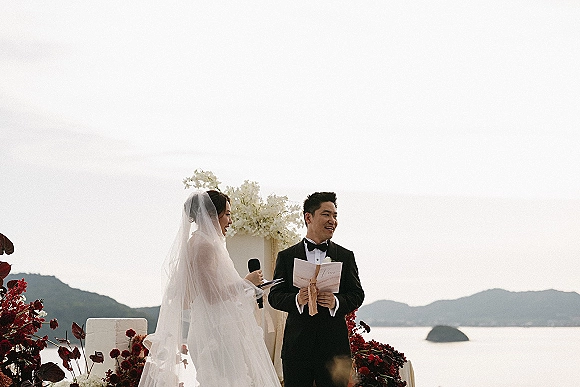 Ceremony moment as bride in veil reads into a handheld microphone beside groom in black tuxedo on a lakeside mountain platform