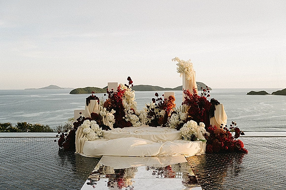 Ceremony altar decor with wedding altar flowers on plinths and draped fabric, mirrored aisle runner and floor cushions facing an ocean-view terrace