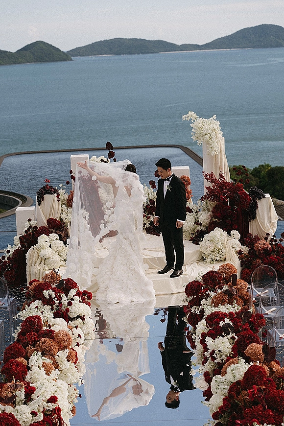 Ceremony setup with mirrored aisle wedding reflections, lined with burgundy and white florals and draped pillars on an ocean-view terrace